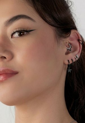 Frauengesicht mit Nahaufnahme eines Ohrs mit fünf schwarzen und silbernen Ohrringen, darunter Stern- und Kreoldesigns, sowie winged eyeliner.