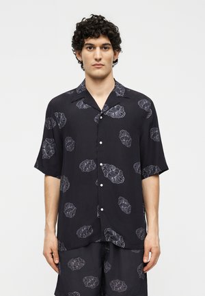 Homme aux cheveux bouclés portant une chemise noire à manches courtes boutonnée et un short assorti avec un motif floral léger, debout devant un fond uni.