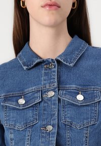 Chaqueta de mezclilla en azul medio con cuello, botones a presión, dos bolsillos en el pecho y detalles de costura vertical. Textura suave.