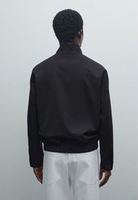 Man wearing a black jacket and white pants stands facing a plain light gray background, shown from the back.