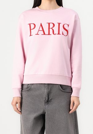 Person wearing a light pink sweatshirt with "PARIS" in large red letters and loose-fitting dark gray jeans, standing against a plain background.