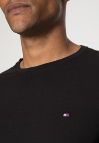Schwarzes Baumwoll-T-Shirt mit Rundhalsausschnitt und einem kleinen roten, weißen und blauen Logo auf der linken Brust. Weiches Material, normale Passform.