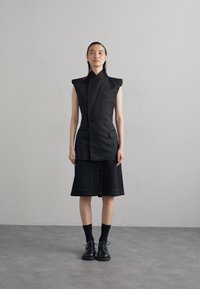 Čierny štruktúrovaný krátky top bez rukávov s vysokým golierom, kombinovaný s šortkami po kolená. Podrobnosti zahŕňajú vrecká a kontrastné šitie. Čierna obuv dotvára celý vzhľad.
