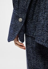 Blazer azul marinho texturizado com um padrão tecido notável, apresentando um botão prateado na manga e um corte elegante e ajustado.