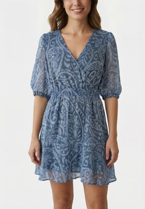 Mujer con vestido cruzado azul con estampado, mangas abullonadas transparentes y cintura elástica, posando frente a un fondo blanco liso.