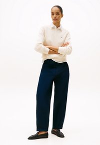Femme debout, bras croisés, portant un pull texturé blanc, une chemise blanche, un pantalon bleu marine et des mocassins noirs sur un fond uni.