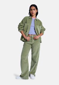 Πράσινο oversized σακάκι από κορδούρα με δύο τσέπες μπροστά, συνδυασμένο με μπλε και λευκό ριγέ κροπ τοπ και πράσινο παντελόνι με φαρδιά μπατζάκια. Λευκά αθλητικά παπούτσια.