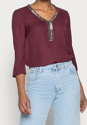 Kobieta w bordowej bluzce z dekoltem w serek wykończonej metaliczną lamówką oraz jasnoniebieskich, wysokich dżinsach, stojąca z opuszczonymi rękami.