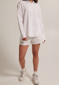 Osoba v nadměrné bílé košili na knoflíky, bílých šortkách, bílých ponožkách a bílých teniskách na neutrálním pozadí.