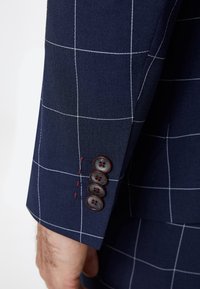 Navy blazer with a plaid pattern; features red stitching and four dark buttons on the cuff; smooth, textured fabric.