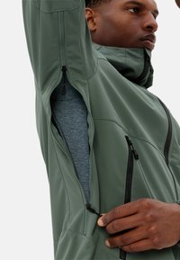 Grüne, leichte Jacke mit hohem Kragen, Reißverschlusstaschen und detaillierten Nähten; getragen über einem grauen Kleidungsstück mit einer glatten Textur.