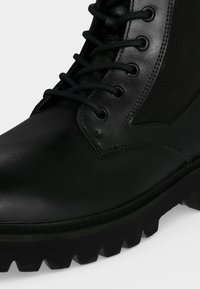 Botte noire en cuir à lacets, avec semelle épaisse striée et design montant à la cheville, montrant des coutures détaillées près des œillets et une finition lisse.