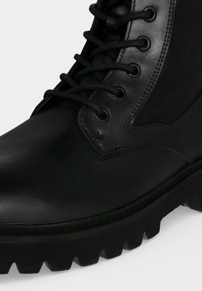 Botte noire en cuir à lacets, avec semelle épaisse striée et design montant à la cheville, montrant des coutures détaillées près des œillets et une finition lisse.