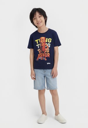 Garçon souriant portant un t-shirt bleu marine avec un personnage de batte de baseball dessin animé et un short en denim, debout devant un fond uni.