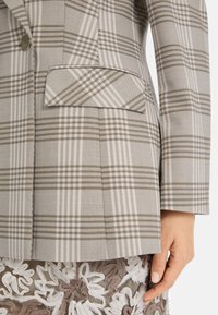 Grauer und beiger Karoblazer mit einer Brusttasche und einem strukturierten Design. Glatte Textur mit taillierter Passform und subtilen Musterdetails.