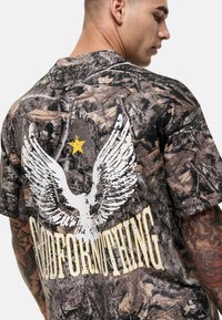 Koszulka w kamuflażowym wzorze z białym grafiką orła i żółtym napisem "GOODFORNOTHING" na plecach. Wykonana z miękkiego, lekkiego materiału.