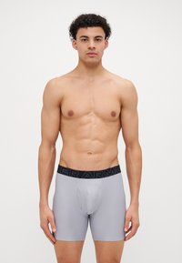 Boxerii gri, dintr-un material elastic și neted, cu un brâu negru inscripționat cu "UNDER ARMOUR". Au un pachet conturat și un design ajustat.