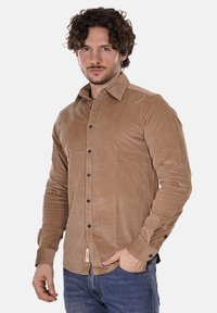 Camicia button-up marrone chiaro a maniche lunghe con una consistenza morbida, bottoni neri e colletto classico. Indossata con jeans blu.