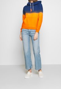 Žena v modro-oranžovém gradientovém mikině, světlých modrých rovných džínech a bílých teniskách stojí proti jednobarevnému pozadí.