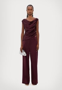 Burgundy draperad topp i kombination med vida burgundy byxor. Modellen håller en silverrandig clutch. Slätt tyg med en subtil lyster.