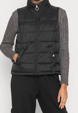 Osoba nosząca czarną pikowaną kamizelkę bez rękawów z suwakiem na pierścień oraz szary, prążkowany sweter z długim rękawem, jedną ręką w kieszeni kamizelki.