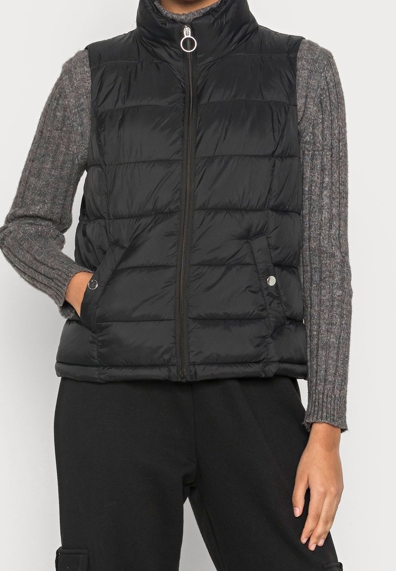Personne portant un gilet matelassé noir sans manches avec une fermeture éclair à anneau et un pull gris à manches longues côtelé, une main dans la poche du gilet.