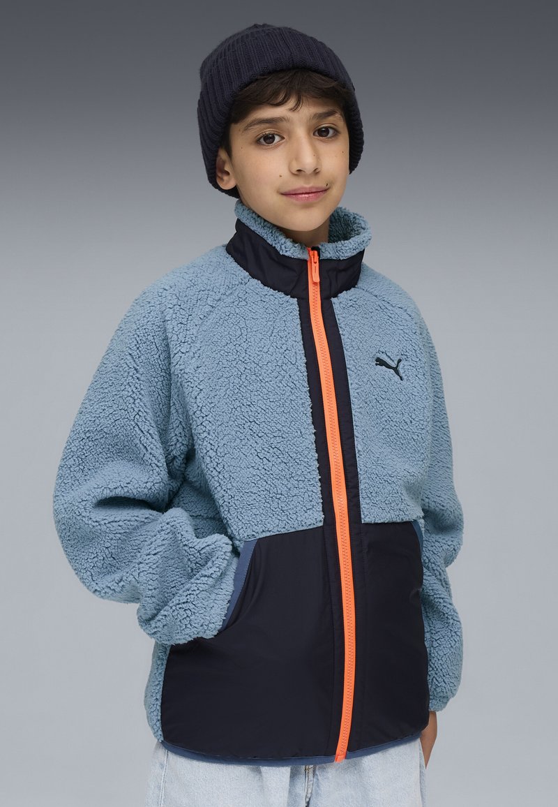 Ljusblå fleecejacka med hög krage, orange dragkedja och marinblå detaljer. Har sidofickor och en texturerad yta.