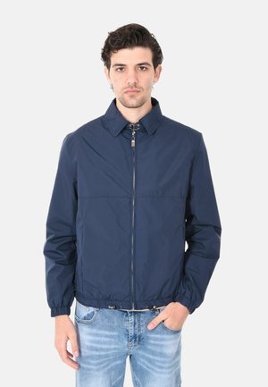 Giovane uomo con giacca con cerniera blu navy e polsini elastici e jeans azzurri, in piedi davanti a uno sfondo chiaro e uniforme.