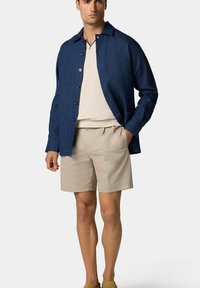 Blaues Leinenhemd mit Knopfleiste, zwei Brusttaschen, über einem cremefarbenen V-Ausschnittshirt und beigen Shorts getragen, kombiniert mit braunen Slippern.