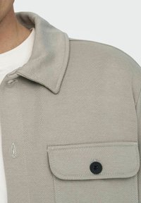 Gray textured jacket with a pointed collar, chest pocket, and black button detail. Fabric features a subtle ribbed pattern.
