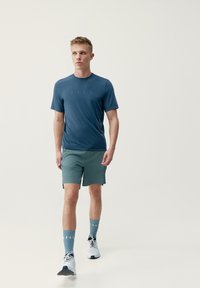 Sportovní outfit se modrým tričkem s krátkým rukávem a zelenými šortkami, doplněný modrými ponožkami a bílými běžeckými botami. Jednoduchý design a střih.