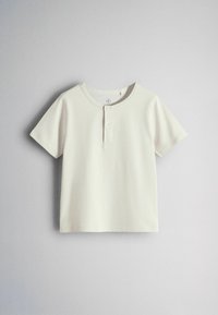Chemise manches courtes de couleur crème avec deux boutons à l'avant et un col rond, présentée sur un fond gris clair.