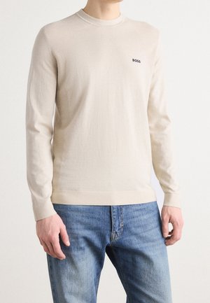 Homme portant un pull beige à col rond avec le logo "BOSS" sur la poitrine et un jean bleu, debout sur un fond blanc uni.