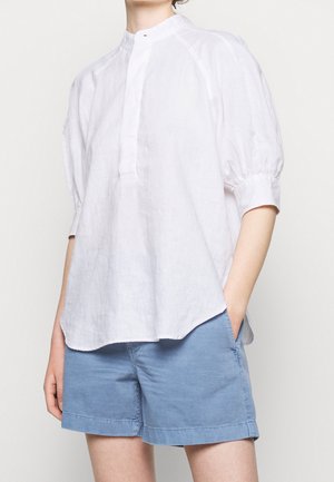 Osoba w luźnej białej bluzce z bufkami na rękawach i niebieskich casualowych szortach, jedną ręką w kieszeni, stojąca na jednolitym tle.