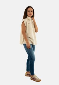 Top beige senza maniche con texture a righe verticali, abbinato a jeans skinny blu e sneakers multicolori. In piedi contro uno sfondo semplice.