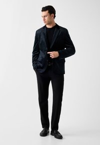 Zwarte fluwelen blazer met een slim fit, twee voorzakken, ingesneden revers en lange mouwen, gecombineerd met zwarte broek en schoenen.