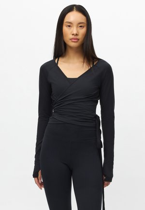 Frau mit langen schwarzen Haaren, die ein figurbetontes schwarzes Wickeloberteil und schwarze Leggings trägt, steht vor einem schlichten weißen Hintergrund.