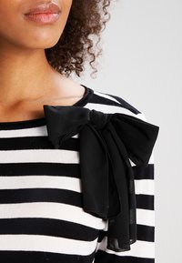 Pull à rayures noires et blanches avec un grand nœud en chiffon noir accentuant l'épaule gauche. Texture douce et coupe décontractée.