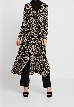 Nainen, jolla on päällään leopardi-kuvioinen pitkä kietaisumekko mustan poolopaidan ja mustien, leveälahkeisten housujen päällä, mustat avojalkaiset korkokengät, taustalla yksivärinen pinta.