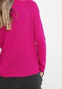 Personne aux cheveux longs châtain clair portant un pull en tricot rose vif et un pantalon noir, vue de dos sur un fond blanc.