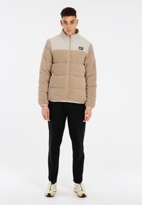 Veste en polaire beige avec une section supérieure gris clair, dotée d'un col montant, d'une fermeture éclair et d'une texture matelassée horizontale. Pantalons noirs et baskets.
