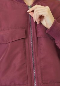 Burgundy bomber jacket with two front pockets, a metal zipper, and ribbed cuffs. Smooth fabric texture with a classic fit.