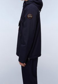 Chaqueta con capucha de color azul marino, fabricada en tejido suave, que presenta un parche de logo en el hombro izquierdo, bolsillos laterales y un cierre de cremallera en la parte inferior.