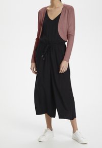 Czarny jumpsuit z dekoltem w serek i ściągaczem w pasie, w połączeniu z jasnoróżowym krótkim kardiganem. Białe sneakersy dopełniają stylizację.