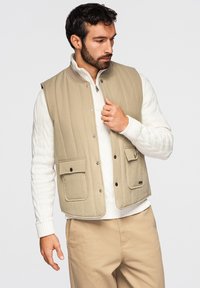 Hombre con barba ajustándose un chaleco beige acolchado sobre un suéter blanco de punto trenzado, vistiendo pantalones beige a juego, de pie frente a un fondo neutro.