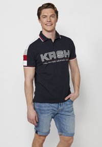 Haditengerészeti póló, kontrasztos szürke és piros ujjakkal, a mellkasán "KRSH" logóval. Világos farmer rövidnadrággal párosítva.