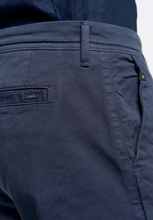 Tumedate sinised chino püksid, mis on valmistatud puuvillase kangast, millel on sile tekstuur. Omab eesmine ja tagumine taskut, vöökohtade silmuseid ning väikest kaubamärgiga sildit.