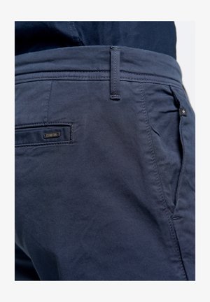 Tumedate sinised chino püksid, mis on valmistatud puuvillase kangast, millel on sile tekstuur. Omab eesmine ja tagumine taskut, vöökohtade silmuseid ning väikest kaubamärgiga sildit.