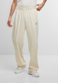 Pantalones de chándal de color crema, de corte holgado, hechos de una tela suave con una cintura elástica. Presenta un pequeño detalle de logo en el muslo izquierdo.