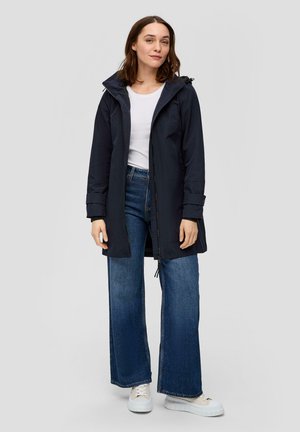 Veste imperméable bleu marine avec capuche, fermeture éclair et poignets ajustables, associée à un jean en denim à jambe large et des baskets blanches.
